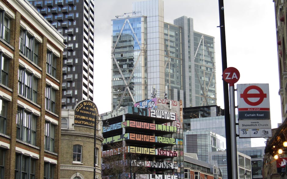 An urban street scene featuring a street sign attached to a pole indicating bus stop information and directions towards Shoreditch Church, with the background showcasing modern glass skyscrapers and o